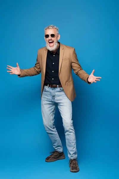 Longitud completa de hombre de mediana edad excitado en gafas de sol gestos en azul - foto de stock