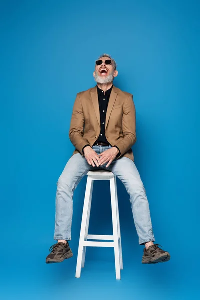 Longitud completa de hombre de mediana edad en gafas de sol riendo en silla blanca en azul - foto de stock