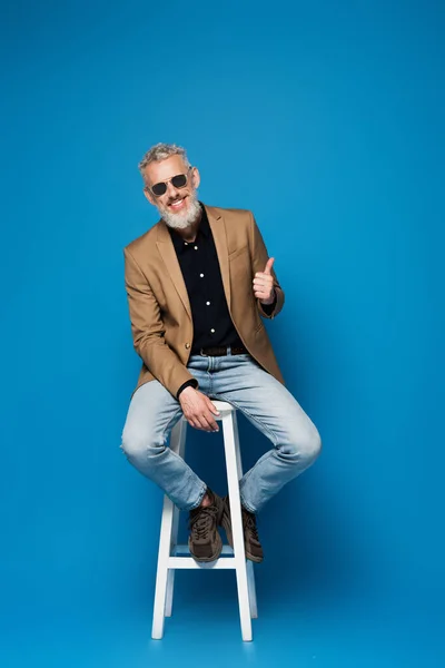 Comprimento total do homem feliz de meia-idade em óculos de sol mostrando polegar para cima enquanto sentado na cadeira branca em azul — Fotografia de Stock