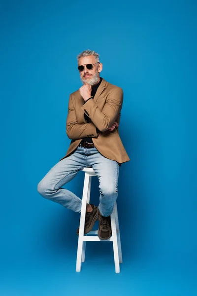 Повнометражний чоловік середнього віку в сонцезахисних окулярах позує на білому стільці на синьому — стокове фото