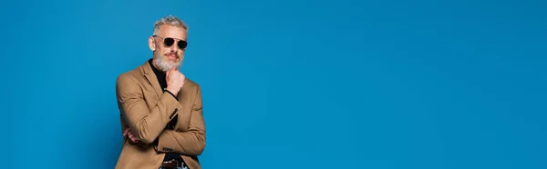 Pensativo hombre de mediana edad en gafas de sol posando aislado en azul, bandera - foto de stock