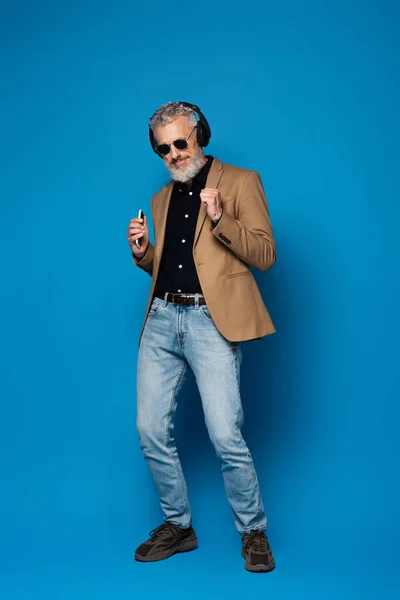 Повна довжина приємного чоловіка середнього віку в сонцезахисних окулярах, що тримає смартфон і слухає музику в бездротових навушниках на синьому — стокове фото