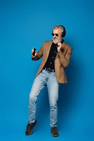 Longitud completa de hombre de mediana edad feliz en gafas de sol celebración de teléfono inteligente y escuchar música en auriculares inalámbricos en azul - foto de stock