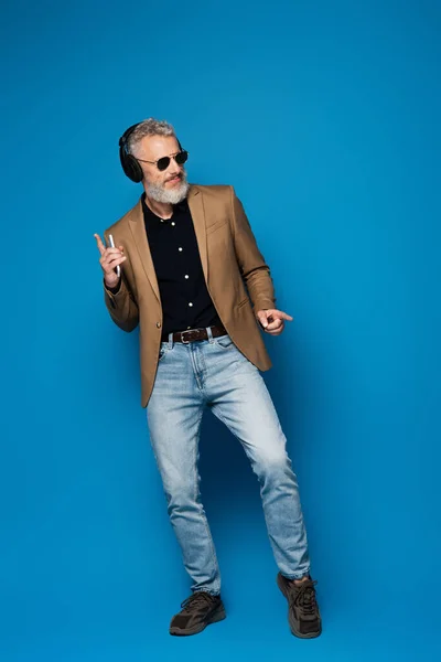 Full length of happy middle aged man in sunglasses holding smartphone and listening music in headphones on blue — Stock Photo