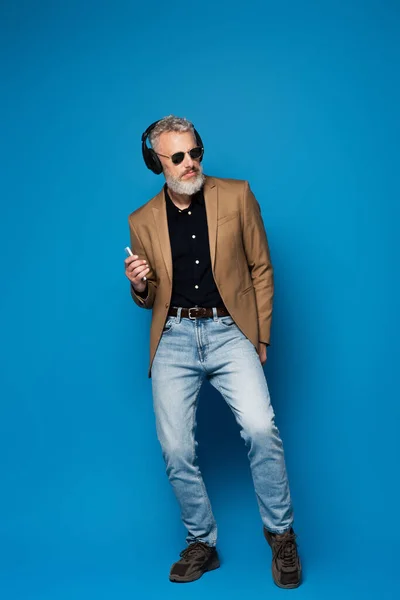 Full length of middle aged man in sunglasses holding smartphone and listening music in headphones on blue — Stock Photo