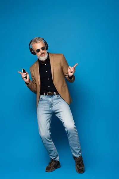 Pleine longueur de sourire homme d'âge moyen dans les lunettes de soleil tenant smartphone tout en écoutant de la musique dans les écouteurs sur bleu — Photo de stock