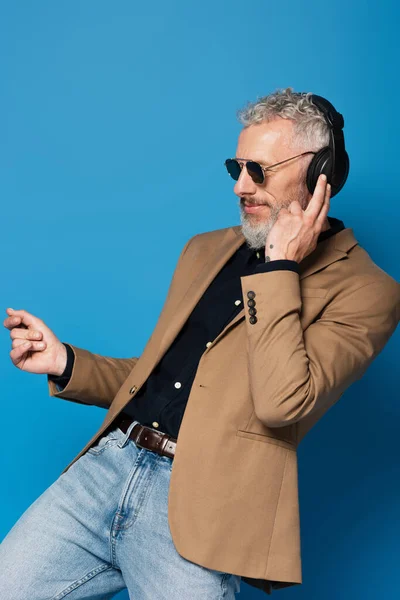 Усміхнений чоловік середнього віку в сонцезахисних окулярах слухає музику в навушниках, танцюючи ізольовано на синьому — стокове фото