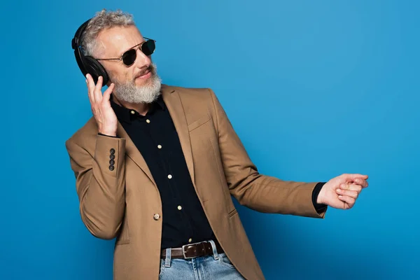 Heureux homme d'âge moyen dans les lunettes de soleil écouter de la musique dans les écouteurs et danser sur le bleu — Photo de stock