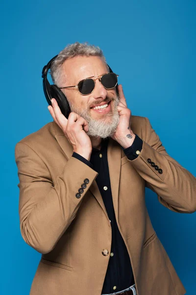 Веселий чоловік середнього віку в сонцезахисних окулярах слухає музику в навушниках ізольовано на синьому — стокове фото