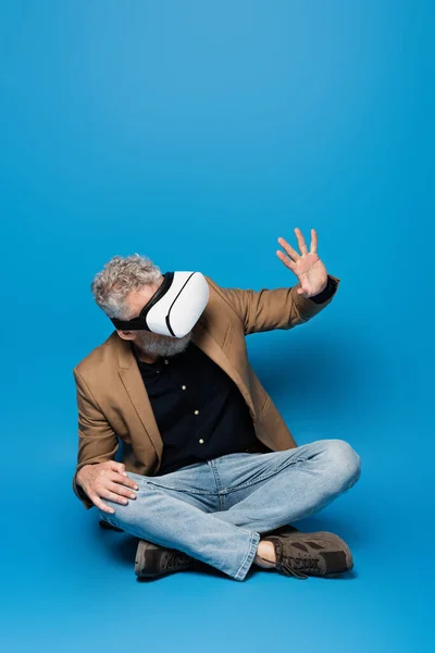 Full length of middle aged man in vr headset sitting with crossed legs and gesturing on blue — Stock Photo