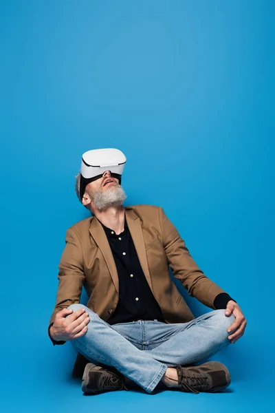 Полный рост мужчина средних лет в гарнитуре VR сидит со скрещенными ногами и смотрит вверх на синем — стоковое фото