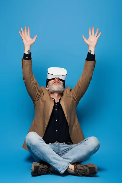 Полный рост мужчина средних лет в гарнитуре VR, сидящий со скрещенными ногами и поднятыми руками на голубом фоне — стоковое фото