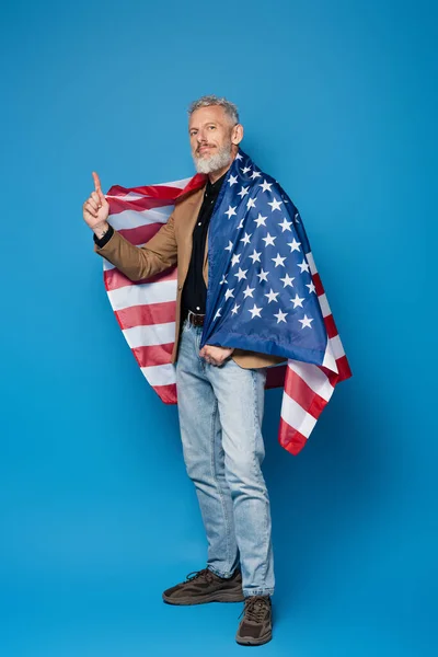 Comprimento total de sorrir homem de meia-idade de pé com bandeira americana e apontando para cima em azul — Fotografia de Stock