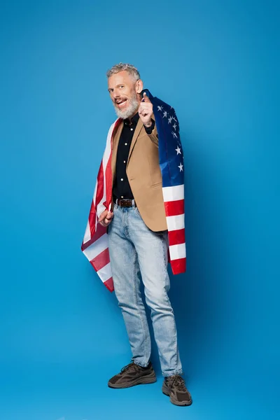 Vollbärtiger Mann mittleren Alters, der mit amerikanischer Flagge auf blauem Hintergrund in die Kamera zeigt — Stockfoto