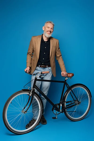 Longitud completa del hombre de mediana edad complacido de pie cerca de la bicicleta en azul - foto de stock