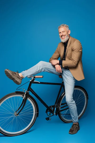 Full length of cheerful middle aged man sitting on bicycle on blue — Stock Photo