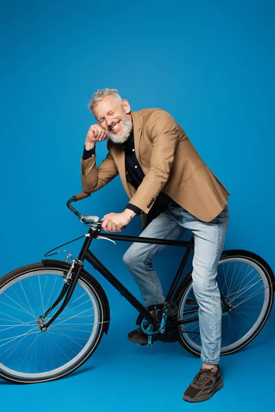 Full length of pleased middle aged man sitting on bicycle and smiling on blue — Stock Photo