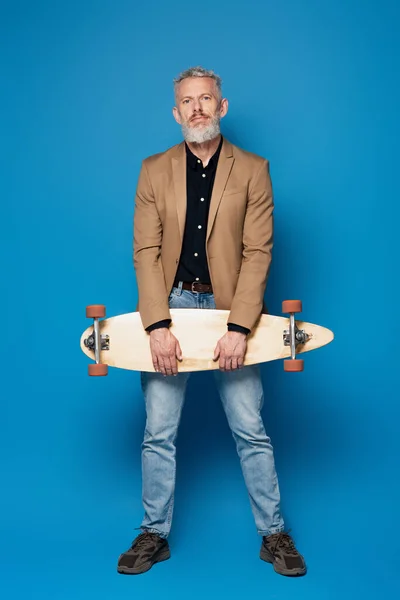 Comprimento total do homem de meia idade barbudo com cabelos grisalhos de pé com longboard em azul — Fotografia de Stock