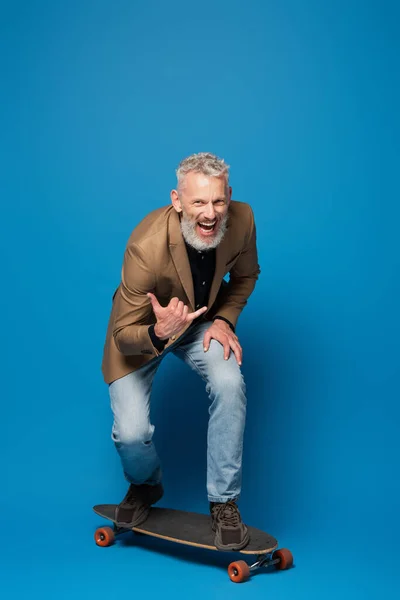 Longitud completa de hombre de mediana edad excitado montando en longboard y gestos en azul - foto de stock