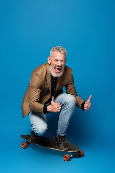 Full length of pleased middle aged man riding on longboard and showing thumbs up on blue — Stock Photo