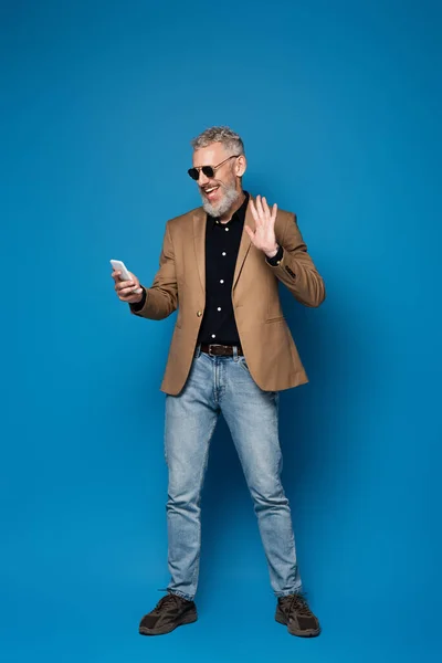 Pleine longueur de heureux homme d'âge moyen dans les lunettes de soleil ayant appel vidéo sur téléphone portable et agitant la main sur le bleu — Photo de stock