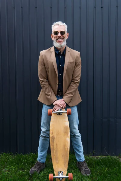 In voller Länge glücklicher Mann mittleren Alters mit Sonnenbrille und Longboard draußen — Stockfoto