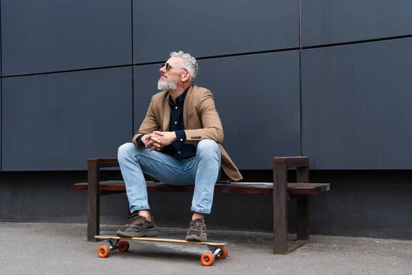 Mann mittleren Alters mit Sonnenbrille sitzt draußen auf Bank neben Longboard — Stockfoto