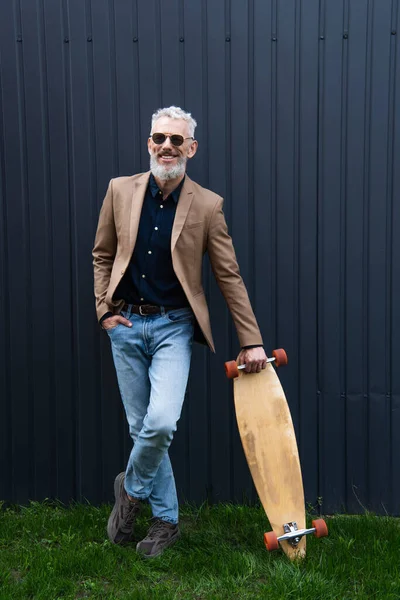 Pleine longueur de heureux homme d'âge moyen dans des lunettes de soleil tenant longboard à l'extérieur — Photo de stock