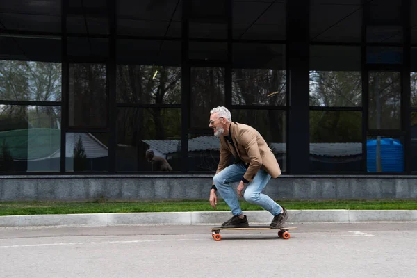 Full length of middle aged man in sunglasses riding longboard near building — Stock Photo