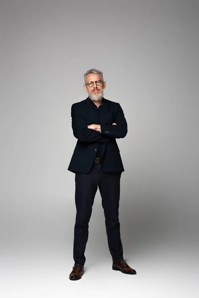 Full length of businessman in glasses and suit posing with crossed arms on grey — Stock Photo