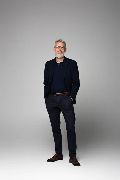 Full length of businessman in glasses and suit posing with hands in pockets on grey — Stock Photo