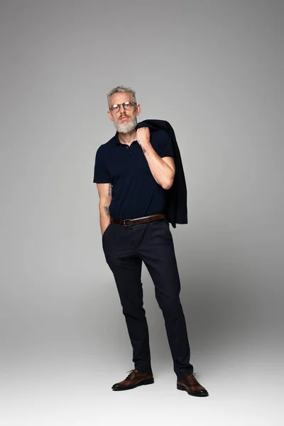 Ganzkörpertätowierter Geschäftsmann in Brille und Anzug posiert mit der Hand in der Tasche auf grau — Stockfoto