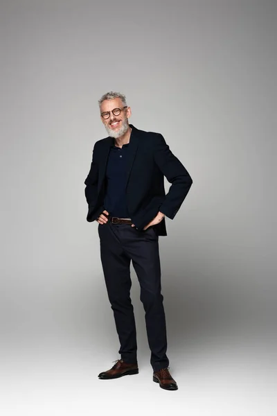 Full length of smiling man in glasses and suit posing on grey — Stock Photo