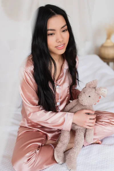 Молода азіатка в рожевій шовковій піжамі сидить з схрещеними ногами на ліжку і тримає м'яку іграшку в спальні — стокове фото
