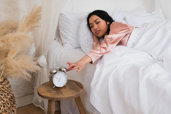 Jeune femme asiatique couché sur du linge blanc et étirant la main à la table de chevet avec réveil — Photo de stock