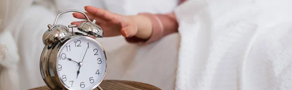 Vista de cerca de la mano femenina extendiéndose a la mesita de noche con reloj despertador en el dormitorio, pancarta - foto de stock