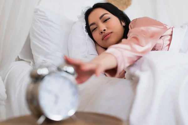 Сонная молодая азиатка лежит в постели и протягивает руку к будильнику на тумбочке в спальне — стоковое фото