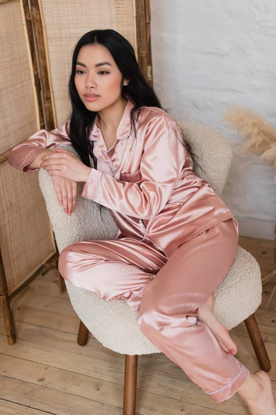 Mujer asiática joven soñadora en pijama de seda rosa sentada en silla en el dormitorio - foto de stock