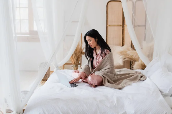Joven mujer asiática en pijama de seda y manta sentado en la cama con las piernas cruzadas y mecanografía en el ordenador portátil en el dormitorio - foto de stock