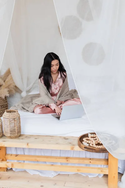 Ernsthafte junge asiatische Frau in seidenem Pyjama und Decke sitzt mit überkreuzten Beinen auf dem Bett und tippt auf dem Laptop im Schlafzimmer — Stockfoto