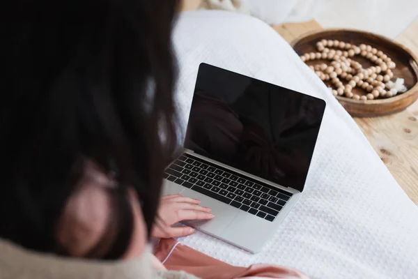 Cropped view of young woman sitting on bed and typing on laptop in bedroom — Stock Photo