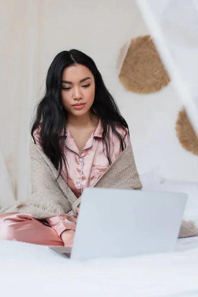 Serious young asian woman in silk pajamas and blanket sitting on bed with crossed legs and working on laptop in bedroom — Stock Photo