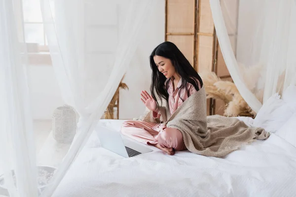 Fröhliche junge asiatische Frau sitzt auf dem Bett und winkt mit der Hand, während sie Videotelefonie mit Laptop im Schlafzimmer hat — Stockfoto