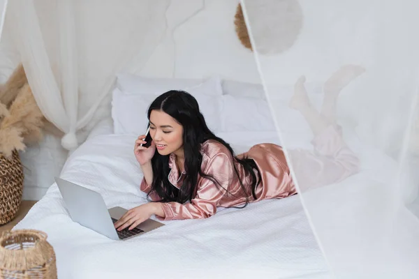 Giovane donna asiatica in pigiama di seta sdraiata sul letto, parlando sul cellulare e guardando il computer portatile in camera da letto — Foto stock