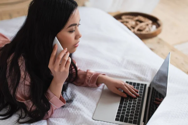 Joven mujer asiática en pijama de seda acostada en la cama, hablando por teléfono celular y mirando a la computadora portátil con pantalla en blanco en el dormitorio - foto de stock