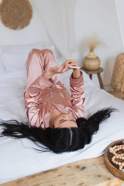 Jeune femme asiatique en pyjama de soie couché sur le lit avec téléphone portable dans la chambre — Photo de stock