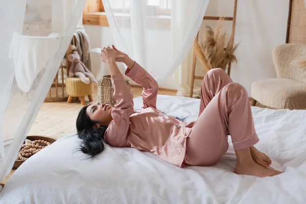 Jeune femme asiatique en pyjama de soie couché sur le lit et tenant le téléphone portable dans les mains dans la chambre — Photo de stock