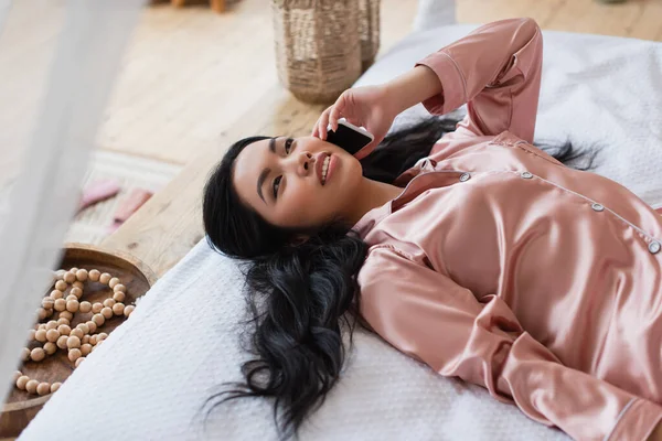 Joven mujer asiática en pijama de seda acostada en la cama y hablando por teléfono celular en el dormitorio - foto de stock