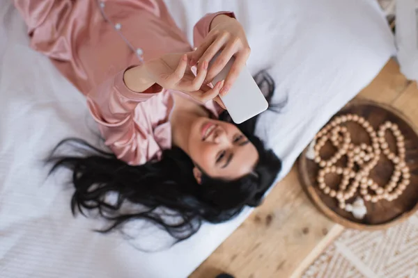 Вид зверху усміхненої молодої азіатки в шовковій піжамі лежить на ліжку і приймає селфі з мобільним телефоном у спальні — стокове фото