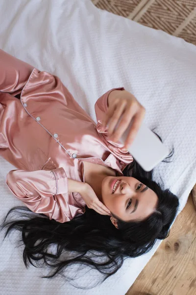 Draufsicht einer jungen Asiatin im seidenen Pyjama, die mit der Hand im Gesicht auf dem Bett liegt und im Schlafzimmer ein Selfie mit dem Handy macht — Stockfoto
